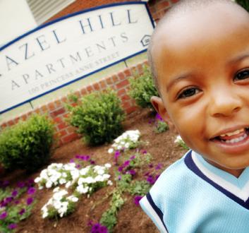 young boy smiling at the camera wearing a light blue vest in front of a sign for Hazel Hill apartments